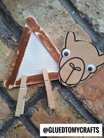 A handmade popsicle stick camel craft featuring a triangular frame, clothespin legs, and a paper camel face with googly eyes, displayed on a brick surface. @GLUEDTOMYCRAFTS is written at the bottom.