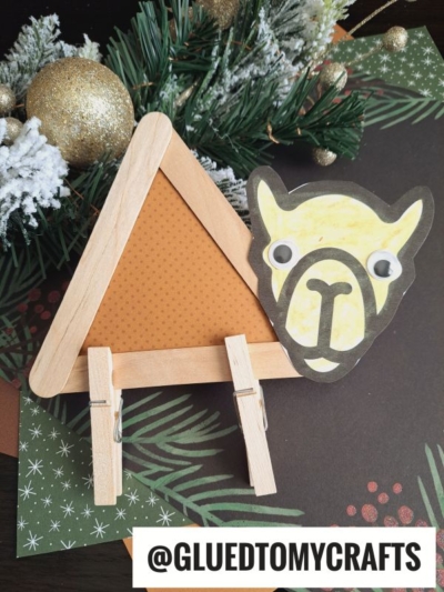 A handmade craft of a lion's face made from paper, googly eyes, and popsicle sticks—much like a popsicle stick camel—with clothespins for legs, displayed on festive paper with a pine branch and ornaments in the background. @GLUEDTOMYCRAFTS text at the bottom.