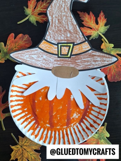 A crafty scarecrow pumpkin gnome made from a paper plate painted orange like a pumpkin, featuring a brown hat, a white handprint beard, and surrounded by paper autumn leaves. @GLUEDTOMYCRAFTS is written at the bottom.