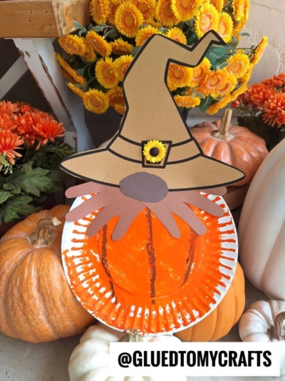 A paper plate craft scarecrow pumpkin gnome with a whimsical witch hat and beard, surrounded by pumpkins and orange flowers outdoors. Text at bottom reads @GLUEDTOMYCRAFTS.