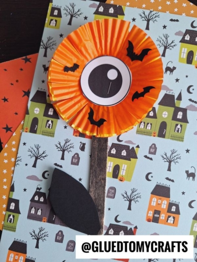 A spooky Halloween craft featuring eyeball flowers made from orange cupcake liners, a large eyeball center, black paper bats, and a wooden stick stem with a black leaf, all set against a festive, eerie background.