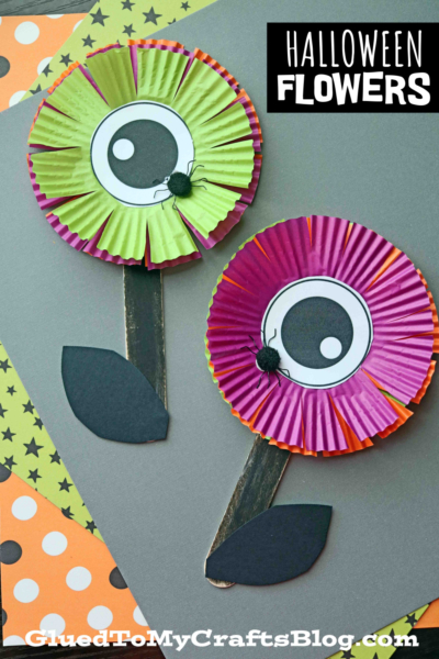 Two Halloween-themed eyeball flowers made from colorful cupcake liners with large cartoon eyes in the center, black paper leaves, and painted stems on a festive background. Text reads Halloween Flowers and GluedToMyCraftsBlog.com.