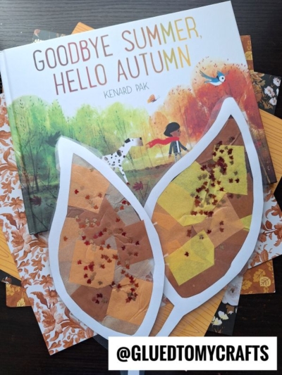 A children’s book titled Goodbye Summer, Hello Autumn lies on patterned paper, with two large leaf suncatcher crafts decorated with colored tissue paper and glitter resting on top. A watermark reads @GLUEDTOMYCRAFTS.