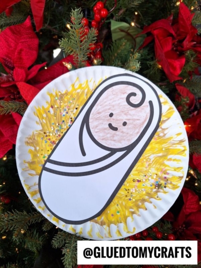 A Paper Plate Manger craft of baby Jesus wrapped in a white blanket, with a smiling face and yellow straw, hangs on a Christmas tree adorned with red poinsettias. @GLUETOMYCRAFTS is written at the bottom.