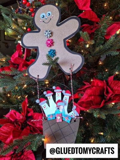 A gingerbread man hot air balloon craft with googly eyes and buttons holds strings attached to a basket filled with Santa figures. The ornament hangs on a Christmas tree decorated with red poinsettias and lights. @GLUEDTOMYCRAFTS appears at the bottom.