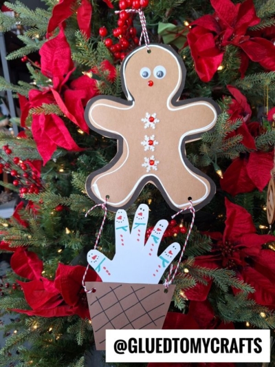 A handmade gingerbread man ornament with googly eyes and buttons hangs on a Christmas tree. Below it, a paper cutout hand, decorated as snowmen, sits inside a “basket” like a gingerbread man hot air balloon. The handle is red and white string.