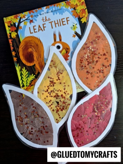 A children’s book titled “The Leaf Thief” with three large, colorful, leaf-shaped crafts resembling a leaf suncatcher, decorated with tissue paper and glitter, displayed on a dark surface. The handle @GLUEDTOMYCRAFTS is at the bottom.