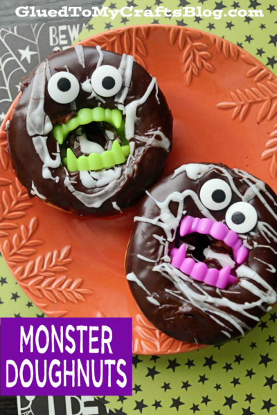 Two chocolate doughnuts with cartoonish monster faces, made with large candy eyes and plastic green and purple fangs, sit on an orange plate. Text reads MONSTER DOUGHNUTS, perfect for fans of playful monster doughnuts.