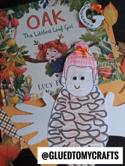 A children’s book titled OAK The Littlest Leaf Girl sits behind a handmade paper craft resembling a pine cone fairy, complete with a smiling face and handprint wings. Autumn-themed decorations surround the items. @GLUEDTOMYCRAFTS.