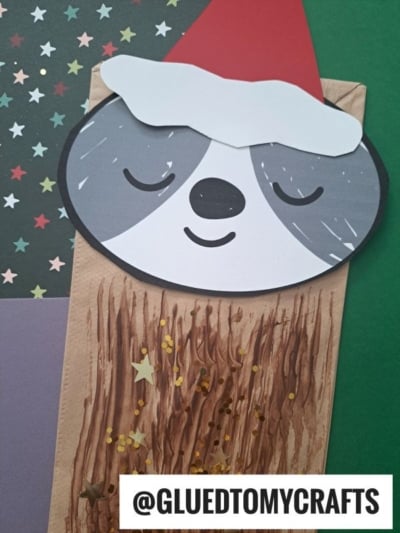 A festive paper bag sloth craft featuring a sloth wearing a red Santa hat, with a painted brown body, gold stars, and a grey and white face, all set against a green background with star-patterned paper nearby.