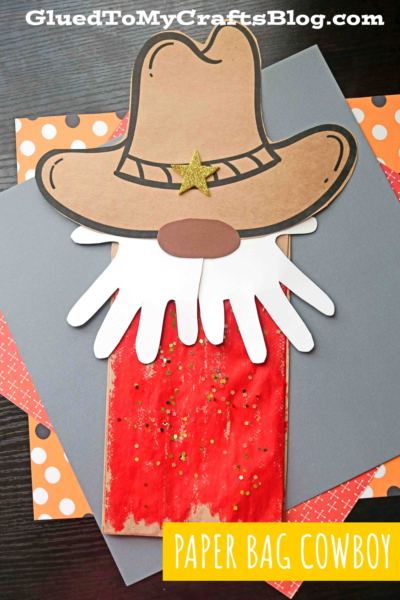 A paper bag cowboy decorated with a large hat, white mustache, red painted shirt, white beard, and gold glitter sits on colored craft paper. The "Paper Bag Cowboy" label and blog watermark are visible.