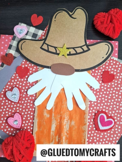 A paper bag cowboy craft featuring a brown paper hat, gold star, white handprint beard, and orange paint, surrounded by red and pink hearts on patterned paper. @GLUEDTOMYCRAFTS is written at the bottom.