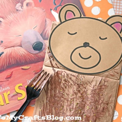 A paper bag sleeping bear craft with a cute bear face is surrounded by colorful paper, a children’s book, and a painted plastic fork.