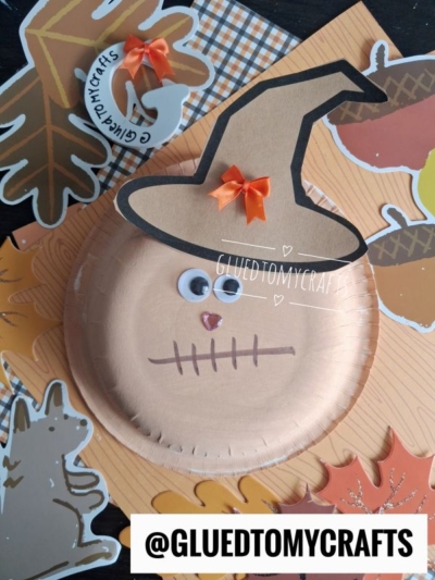 A Paper Bowl Scarecrow with googly eyes, a stitched mouth, and a black hat adorned with an orange bow sits on a table surrounded by autumn-themed paper cutouts.