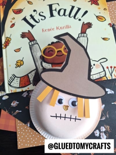 A children’s book titled “It’s Fall!” sits beneath a charming Paper Bowl Scarecrow craft with a brown hat, yellow paper hair, googly eyes, and a stitched mouth. Patterned papers and the @GLUEDTOMYCRAFTS handle are also visible.