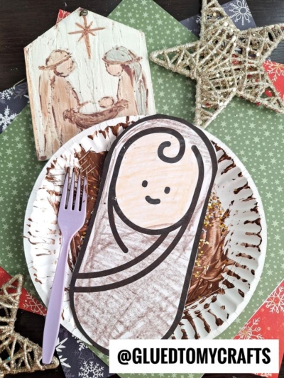 A Paper Plate Manger dessert with chocolate frosting and sprinkles, topped with a paper baby Jesus drawing. The plate sits on festive, star-patterned paper, surrounded by a nativity decoration and gold star ornaments. A purple fork rests beside it.