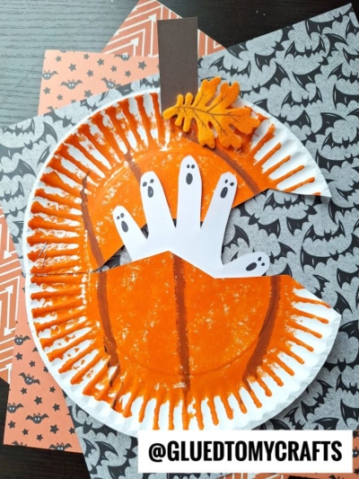 A Halloween craft featuring a paper plate haunted pumpkin, decorated with a handprint cutout of three ghosts, a paper stem, a felt leaf, and layered on bat-patterned paper. @GluedToMyCrafts is labeled below.