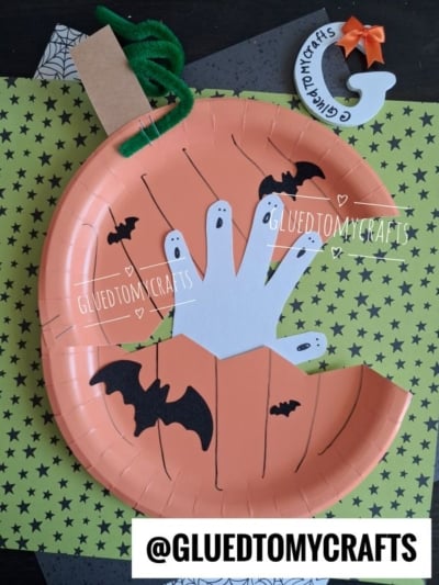 A paper plate haunted pumpkin made from an orange paper plate, decorated with a white handprint ghost, black bats, and a green pipe cleaner stem; Halloween craft supplies are visible.