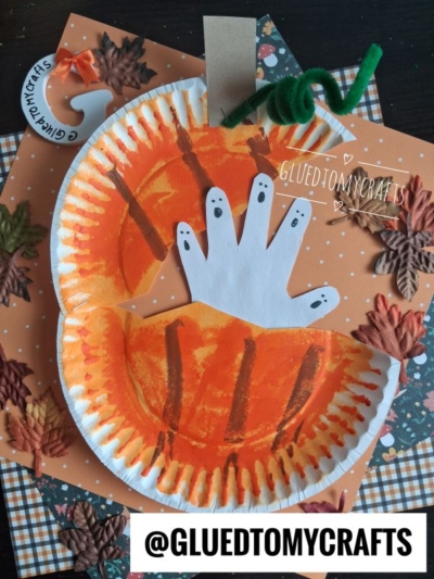 A paper plate haunted pumpkin, painted orange to mimic a festive jack-o’-lantern, features a white handprint with black ghost faces, and is decorated with paper leaves and a curly green stem on a polka-dot background. @GLUEDTOMYCRAFTS is written at the bottom.