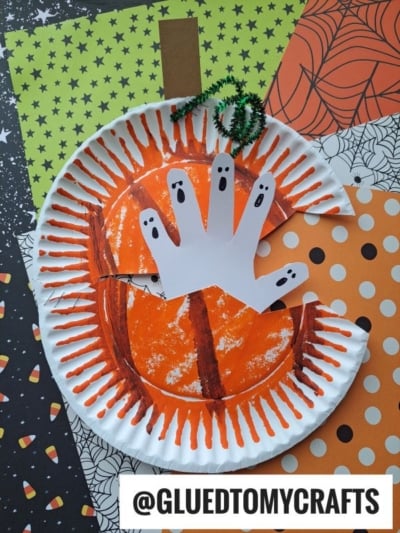 A paper plate haunted pumpkin craft made from a painted paper plate and a cut-out paper hand decorated as ghosts. Halloween-themed papers and a @GLUEDTOMYCRAFTS tag are in the background.
