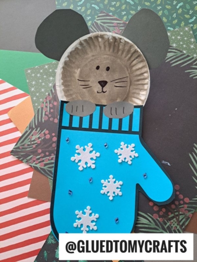 A cute mouse in mitten, crafted from a paper plate and colored paper, with a blue mitten decorated with snowflakes and gems, lies on various patterned craft papers. The handle @GLUEDTOMYCRAFTS is at the bottom.