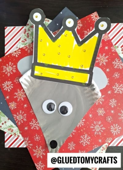 A gray paper plate hedgehog craft with a yellow crown, googly eyes, and a black nose—reminiscent of a whimsical paper plate mouse king—placed on festive red and green snowflake-patterned paper. @GLUEDTOMYCRAFTS text is at the bottom.