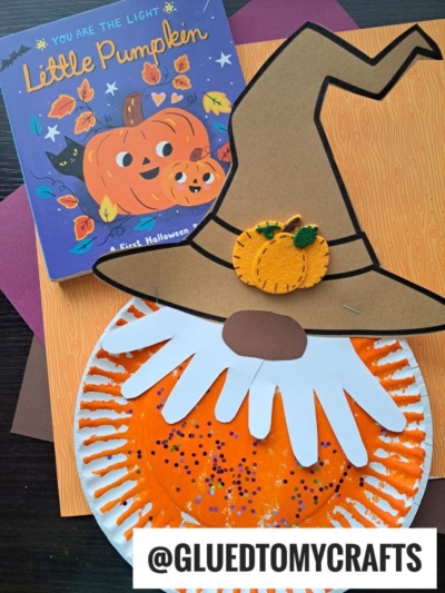 A Halloween craft scene with a pumpkin-themed children’s book, and a paper plate decorated as a scarecrow pumpkin gnome, complete with a felt pumpkin on its hat. The handle @GLUEDTOMYCRAFTS is at the bottom.