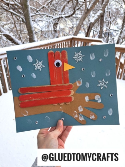 A hand holds a winter-themed craft: a popsicle stick red cardinal made from paper and popsicle sticks, perched on a paper tree branch with snowflakes and finger-painted snow dots on a blue background. Snowy trees and a deck are in the background.