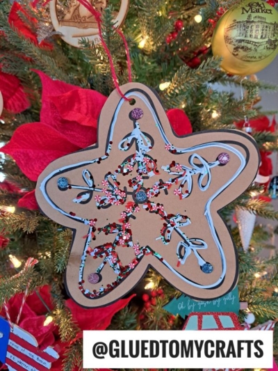 A large, star-shaped cardboard ornament inspired by a gingerbread star cookie ornament is decorated with colorful glitter, paint, and swirl designs. It hangs on a Christmas tree with red poinsettias and ornaments. A tag reads @GLUEDTOMYCRAFTS.