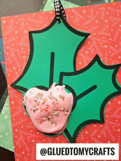 A pink, heart-shaped ornament with festive sprinkles sits on green holly leaf cutouts and red, holiday-patterned paper. Embellished with a black-and-white ribbon, this charming paper mistletoe ornament is labeled @GLUEDTOMYCRAFTS at the bottom.