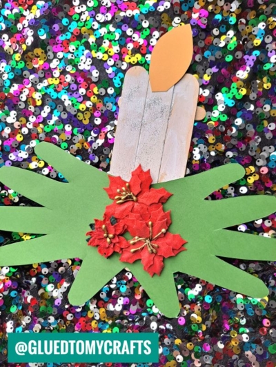 A popsicle stick Christmas candle craft featuring green handprint leaves and red poinsettia flowers, all set against a colorful sequin background.