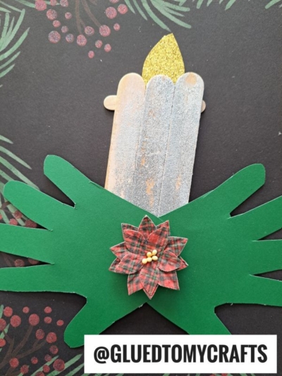A popsicle stick Christmas candle with a yellow flame is wrapped in green paper shaped like hands and adorned with a plaid flower. The dark background features pine and berry patterns. Text reads @GLUETOMYCRAFTS.