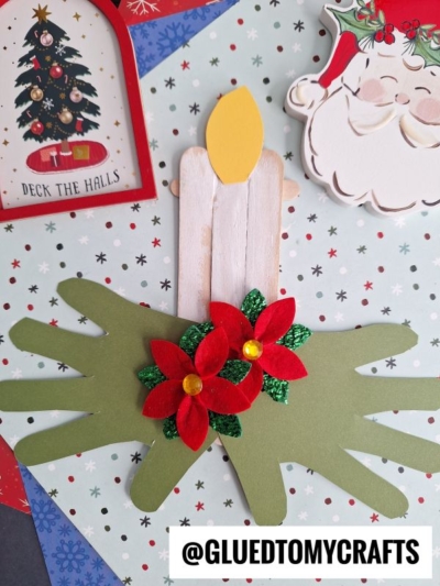 A Christmas craft featuring a popsicle stick Christmas candle with a yellow flame and red poinsettias, set on green paper handprints, surrounded by festive cards and decorations. @GLUEDTOMYCRAFTS text at the bottom.
