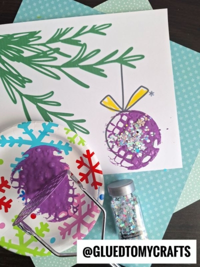 A potato masher stamped Christmas ornament craft features a purple painted ornament with glitter on white paper. A green marker drew a pine branch above, and nearby are a paint roller, purple paint on a snowflake plate, and a jar of glitter.