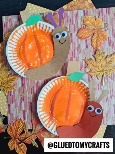 Two paper plate pumpkin snail crafts are decorated as snails with brown and red bodies, googly eyes, and smiling faces. They rest on autumn-themed paper with scattered leaves. @GLUEDTOMYCRAFTS is written at the bottom.