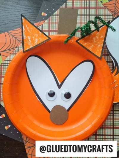 A pumpkin fox face craft made from an orange paper plate, white and black paper for details, googly eyes, and brown and green accents, displayed on a festive background.