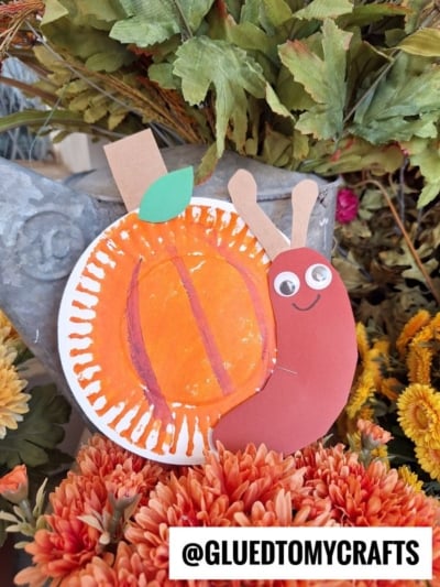 A handmade paper plate pumpkin snail craft with an orange pumpkin shell and googly eyes sits among fall flowers and leaves. @GLUEDTOMYCRAFTS is written at the bottom.