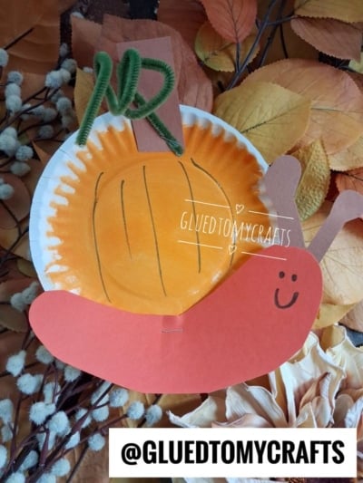 A charming paper plate pumpkin snail craft, featuring a paper plate painted orange to resemble a pumpkin with a green pipe cleaner stem, brown paper stem, and an adorable orange snail body, set atop autumn leaves. GLUEDTOMYCRAFTS watermark and @GLUEDTOMYCRAFTS text included.