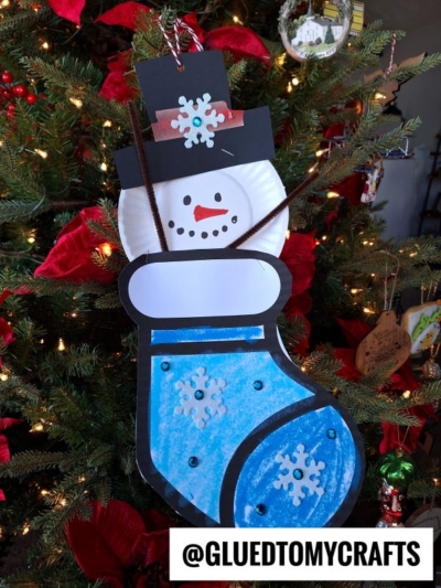 A handmade snowman stocking ornament, shaped like a blue stocking with a snowman’s face in a black hat and decorated with snowflakes, hangs on the Christmas tree. Text at the bottom reads @GLUEDTOMYCRAFTS.
