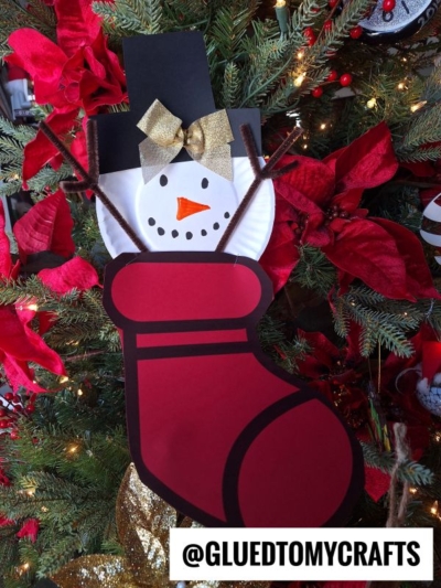 A handmade snowman stocking craft with a paper plate face, gold bow, brown pipe cleaner arms, and a red paper stocking, hanging on a Christmas tree with red poinsettias. @GLUEDTOMYCRAFTS is written at the bottom.