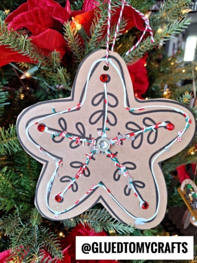 Star-shaped gingerbread star cookie ornament made of brown paper with black doodles, red sequins, white and red twine, and a rhinestone center, hanging on a Christmas tree with red poinsettias. @GLUETOMYCRAFTS at the bottom.