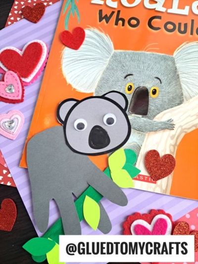 A handmade handprint koala craft with googly eyes sits on a branch over a colorful koala-themed book, surrounded by heart-shaped decorations and felt hearts. The tag @GLUEDTOMYCRAFTS is visible at the bottom.
