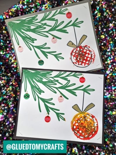 Two festive holiday cards with hand-drawn green pine branches, colorful baubles, and an ornament made of red or yellow glitter on white paper, placed on a sparkly, sequin-filled background. The charming designs are enhanced by potato masher stamped Christmas accents that add playful texture and unique detail to each card.