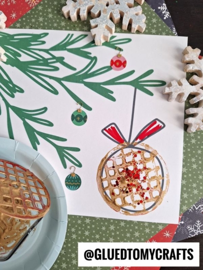A holiday craft scene shows a white paper with painted pine branches, ornament drawings, and a 3D waffle piece with red glitter. Snowflake decorations and festive paper surround this potato masher stamped Christmas artwork. @GLUEDTOMYCRAFTS is printed below.