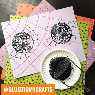 A potato masher stamped craft features two spiders made from black paint and confetti on purple paper with red web lines, set on star-patterned sheets. A paper plate with black paint and a potato masher are nearby. @GLUEDTOMYCRAFTS is labeled.