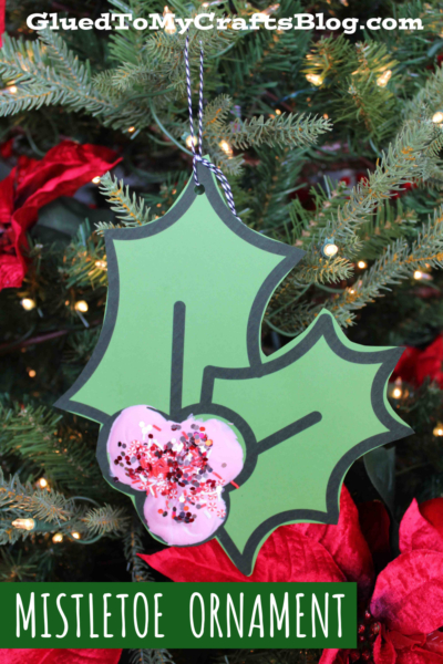 A paper mistletoe ornament with three green leaves and pink berries hangs on a Christmas tree, decorated with red flowers and lights. The image includes GluedToMyCraftsBlog.com, Mistletoe Ornament, and highlights this puffy paint mistletoe craft.