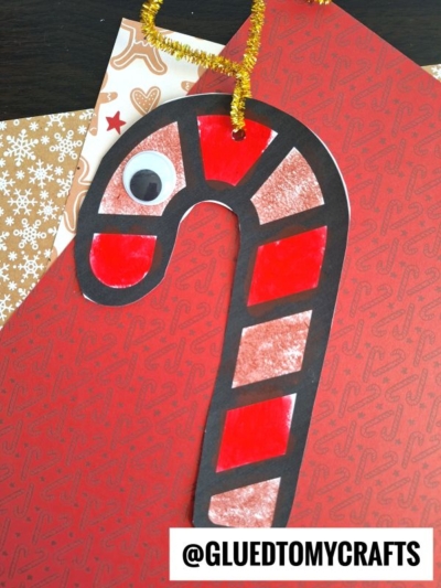 A handmade candy cane reindeer ornament with red and white sections, a black outline, a googly eye, and a gold pipe cleaner loop lies on festive red and snowflake-patterned paper. @GLUEDTOMYCRAFTS is written at the bottom.