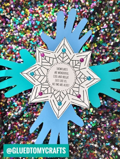 A handmade handprint snowflake craft made of layered blue paper hand shapes and a decorated paper snowflake with colorful gems. The snowflake’s center reads, “Snowflakes are wonderful, cool and bright… Just like us, no two are alike!”.
