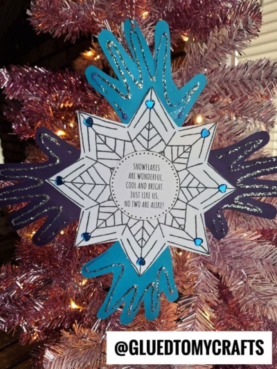 A handmade handprint snowflake ornament made from blue and purple hand-shaped paper cutouts, decorated with blue gems, hangs on a pink Christmas tree. In the center: “Snowflakes are wonderful, cool and bright, just like us! No two are alike!”.