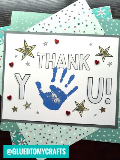 A handmade handprint thank you card with Y, a blue handprint, and U! spelling Thank You! among gold stars and red heart stickers, placed on patterned papers. The handle @GLUEDTOMYCRAFTS appears at the bottom.